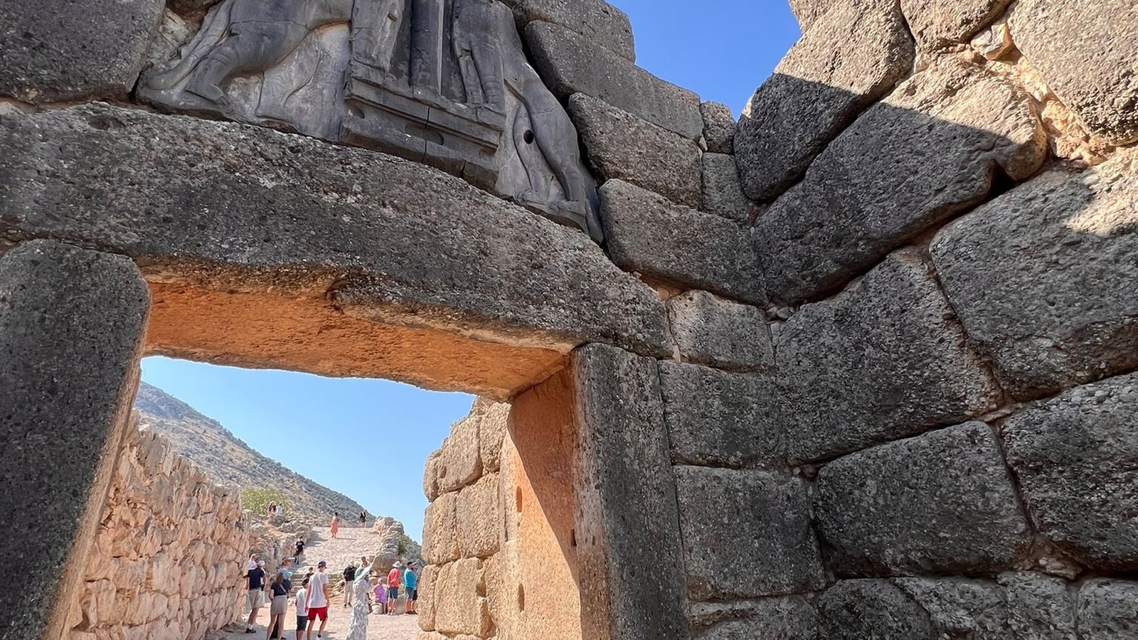 Antikes Steintor aus großen Quaderblöcken mit gemeißeltem Relief über dem Durchgang, Holzsteg davor, Besucher im Hintergrund